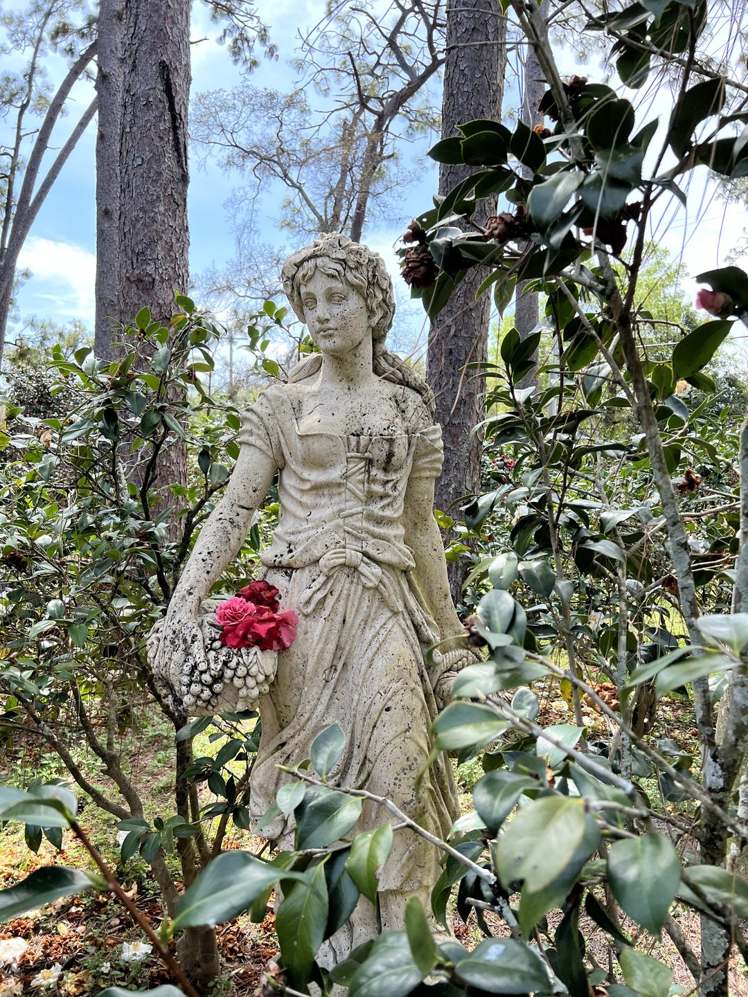 A stone statue of a woman holding flowers stands amidst lush greenery and tall trees, creating a serene garden atmosphere.