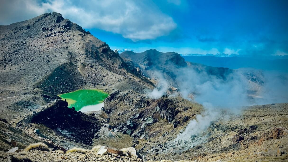 A rugged mountain landscape features a vibrant green lake surrounded by rocky terrain and steam rising from geothermal vents.
