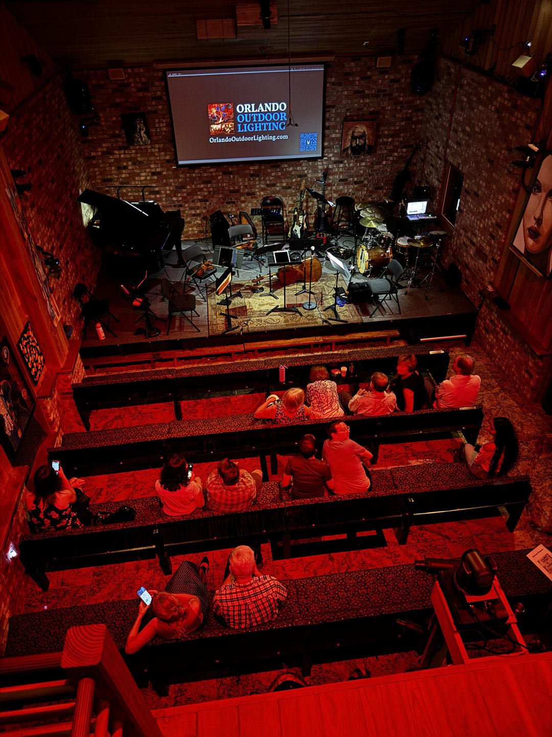 A cozy music venue with wooden benches and a stage set for a performance. Audience members are seated, with instruments visible.