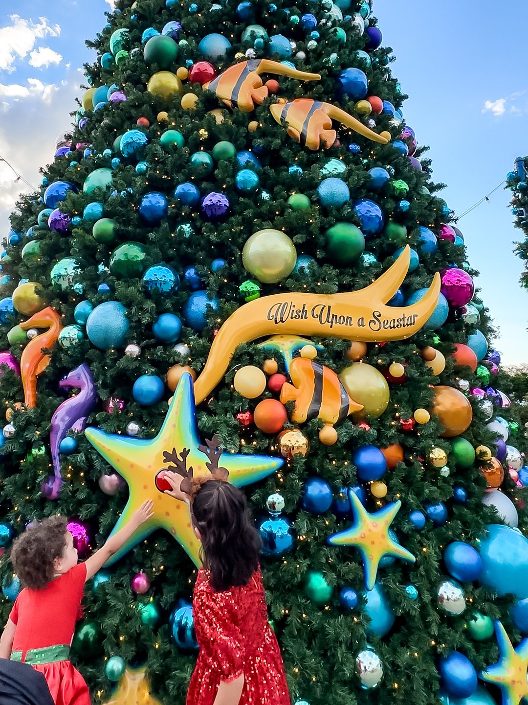 A vibrant Christmas tree adorned with colorful ornaments, featuring playful sea-themed decorations and children reaching for the stars.