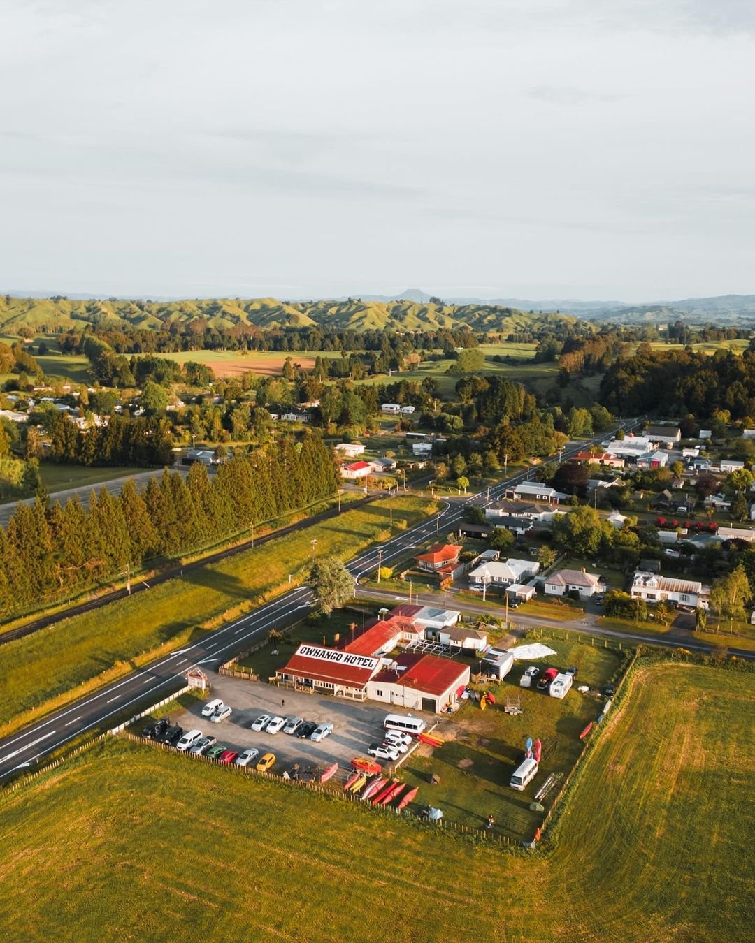 Aerial view of a small town surrounded by green hills, featuring a mix of residential and commercial buildings along a road.