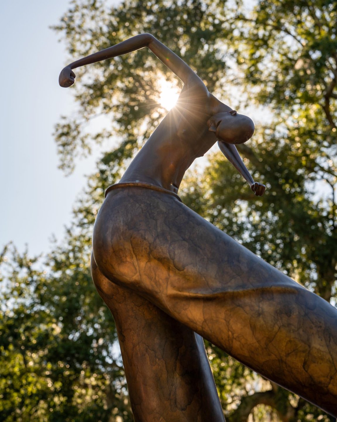 A bronze sculpture of a stylized figure with an elongated neck and arms, backlit by sunlight, surrounded by lush greenery.