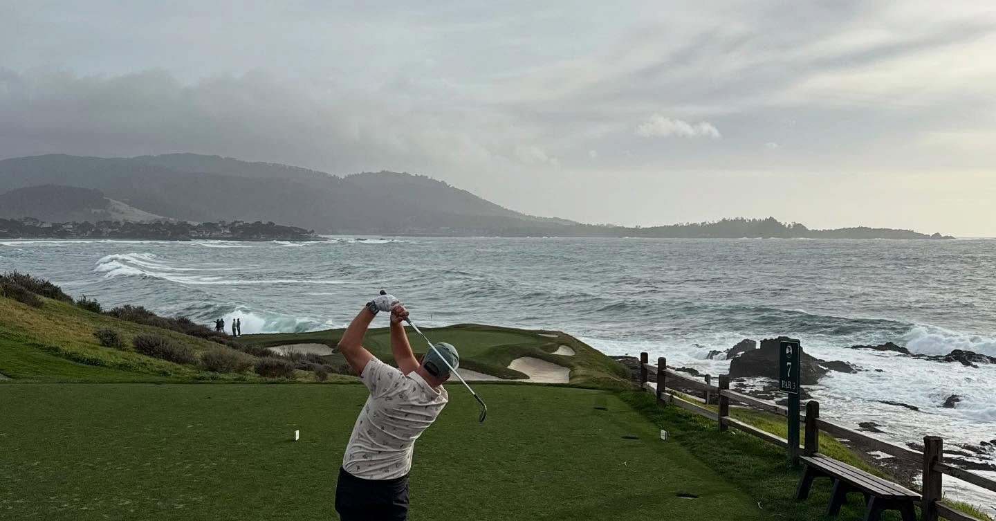 A golfer swings on a coastal course, with waves crashing nearby and rolling hills in the background under a cloudy sky.