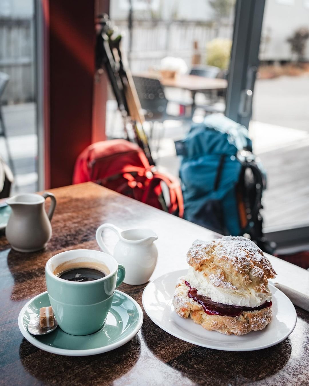 A cozy café scene featuring a cup of coffee and a scone with cream and jam on a table, with colorful backpacks in the background.