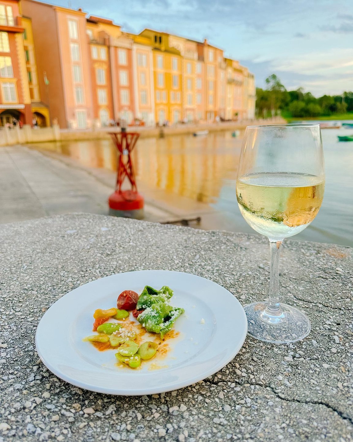 A plate of colorful pasta with vegetables sits on a stone surface, accompanied by a glass of white wine. In the background, vibrant buildings reflect in the water.