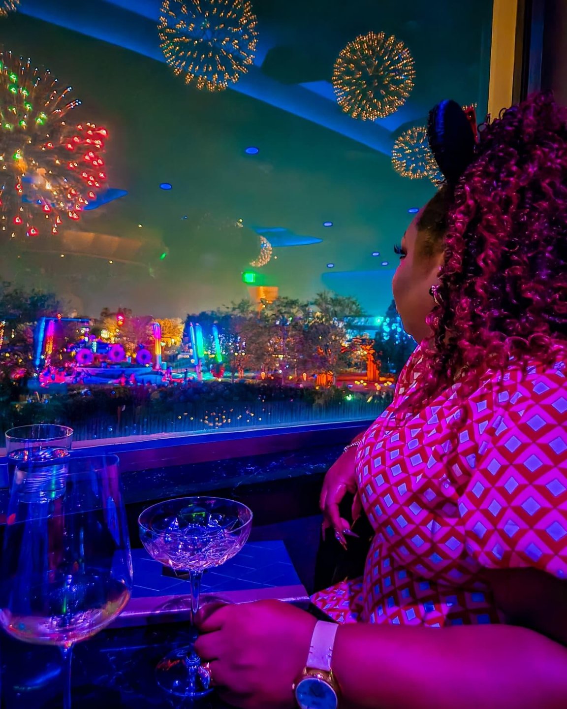 A woman with curly hair sits by a window, watching a vibrant fireworks display over a colorful landscape, with drinks on the table.