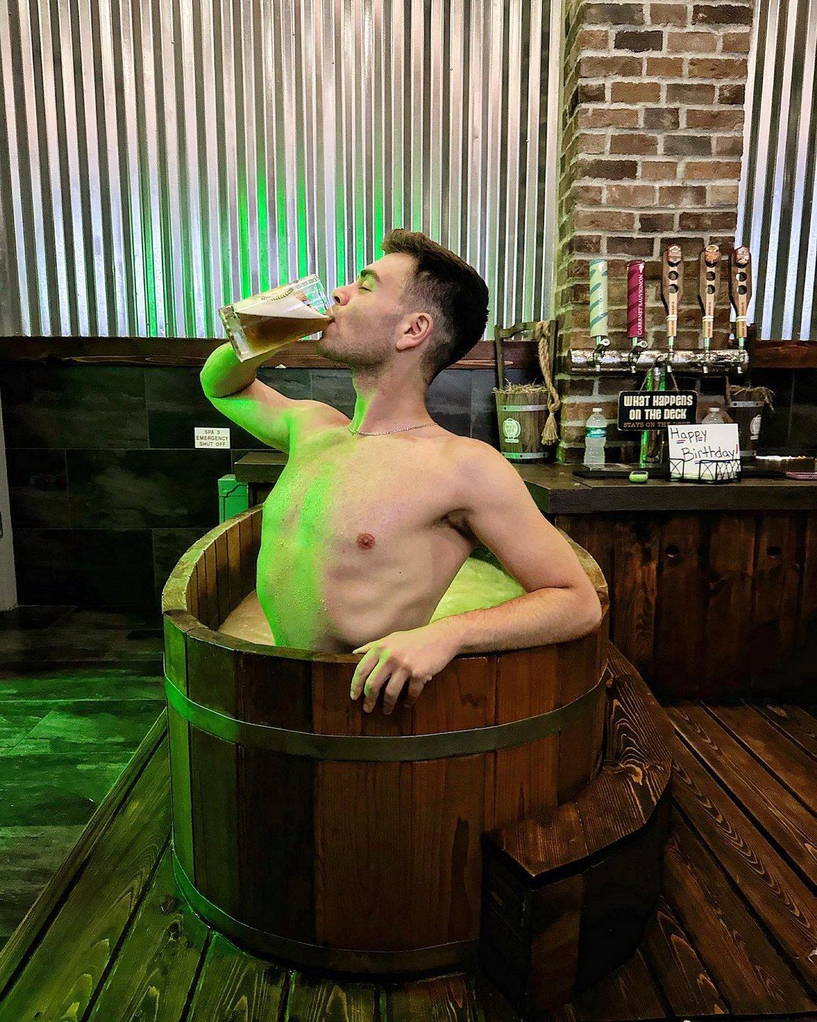 A man sits in a wooden barrel, sipping a drink, surrounded by rustic decor and taps in a cozy, dimly lit setting.