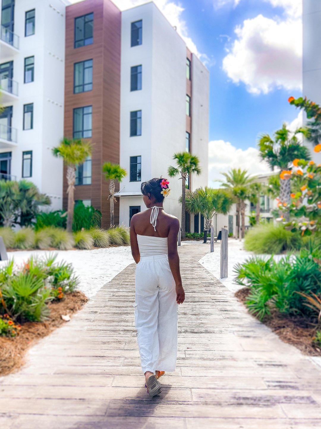 A woman in a white outfit walks along a wooden path surrounded by lush greenery and modern buildings under a bright sky.