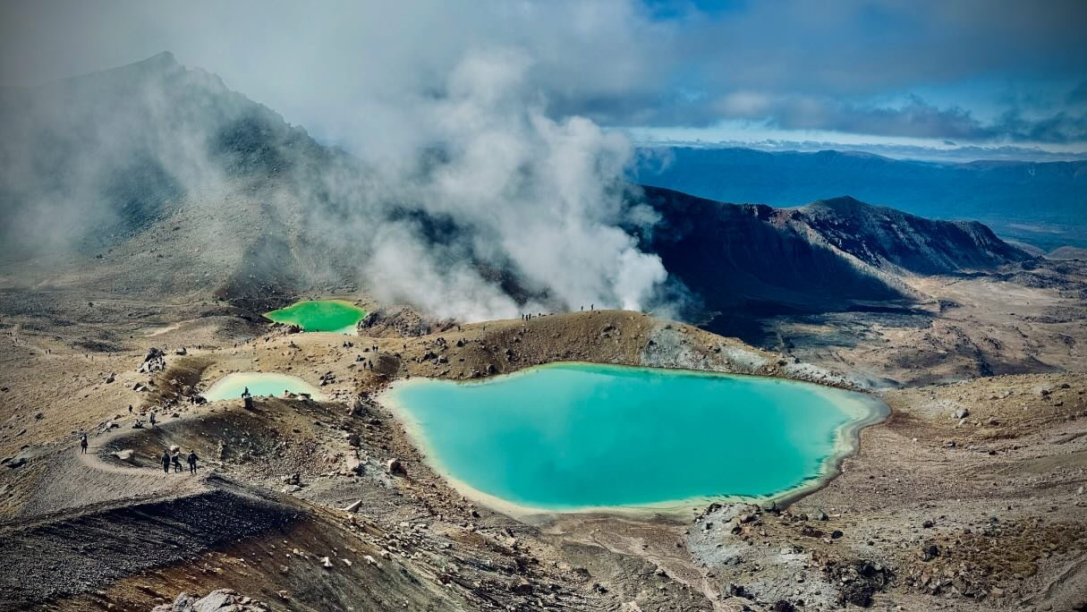 Aerial view of vibrant turquoise lakes surrounded by rugged terrain and misty mountains, with hikers exploring the landscape.