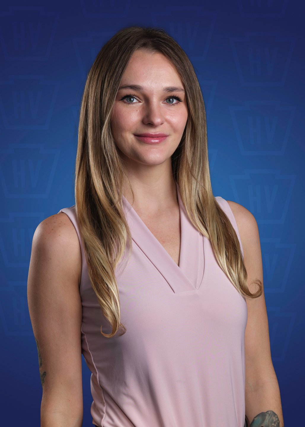 A woman with long hair smiles, wearing a light pink top against a blue background featuring a logo. The image promotes a welcoming atmosphere.