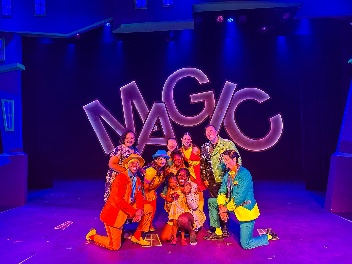 A vibrant stage scene featuring a diverse group of performers in colorful costumes, posing in front of a large "MAGIC" sign under dramatic lighting.