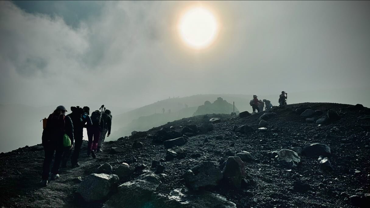 A group of hikers ascends a rocky, mist-covered landscape under a bright sun, with distant hills and boulders visible in the background.