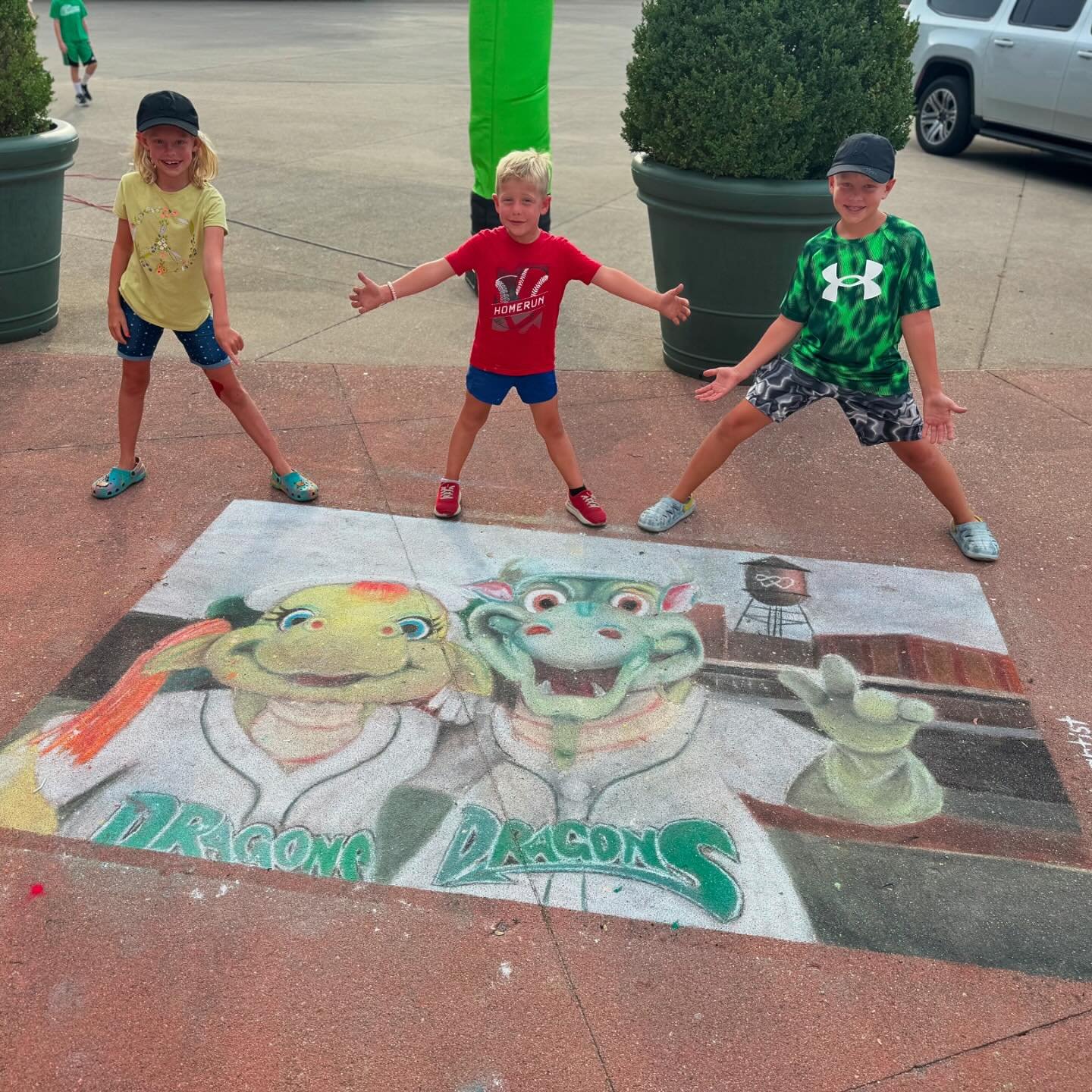 Three children pose playfully around a colorful chalk drawing of cartoon dragons on a pavement. Gree…
