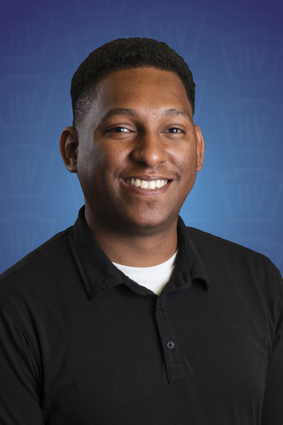 A smiling individual in a black shirt poses against a blue background with a subtle logo pattern, promoting a welcoming atmosphere.