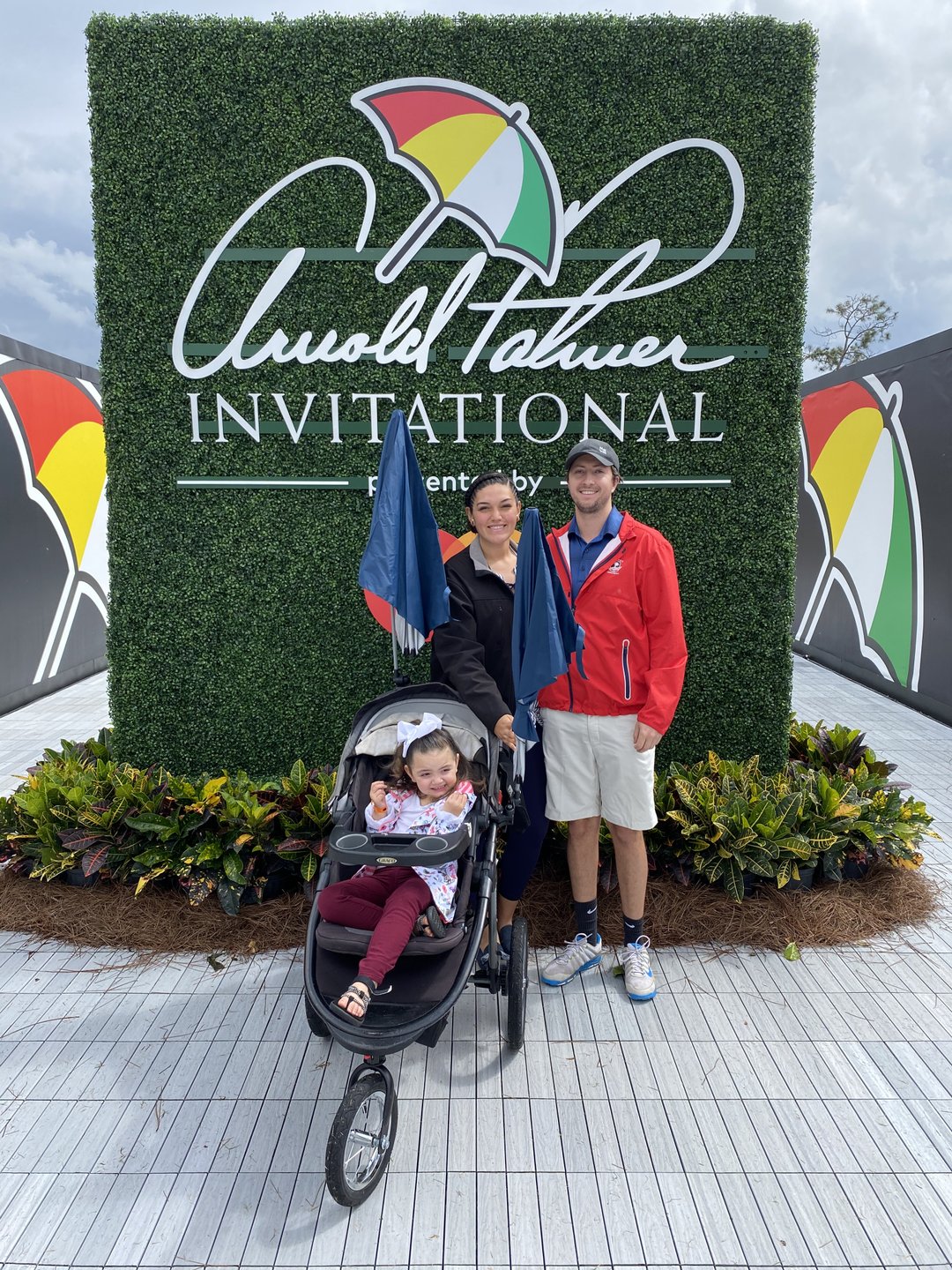 A family poses in front of a large green sign for the Arnold Palmer Invitational, featuring colorful umbrellas. A stroller is nearby.