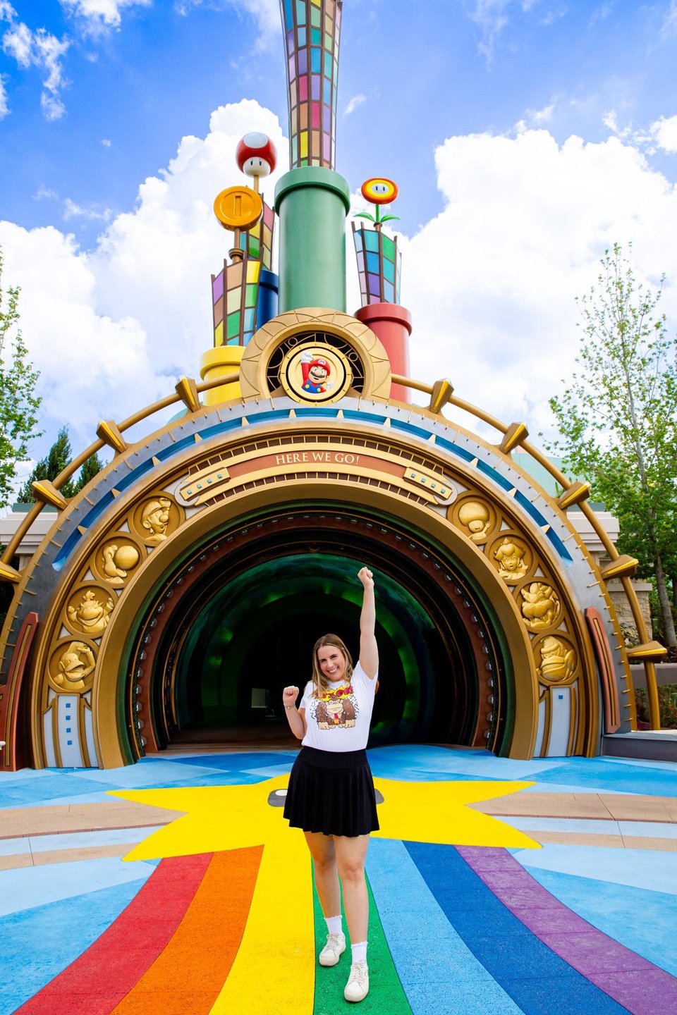 A vibrant entrance with colorful patterns leads into a whimsical attraction. A person stands joyfully in front, raising their arm.