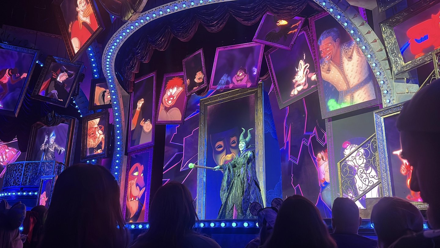 A vibrant stage scene featuring a character in a dramatic costume, surrounded by colorful framed portraits of various animated figures. The audience is visible in the foreground, captivated by the performance.