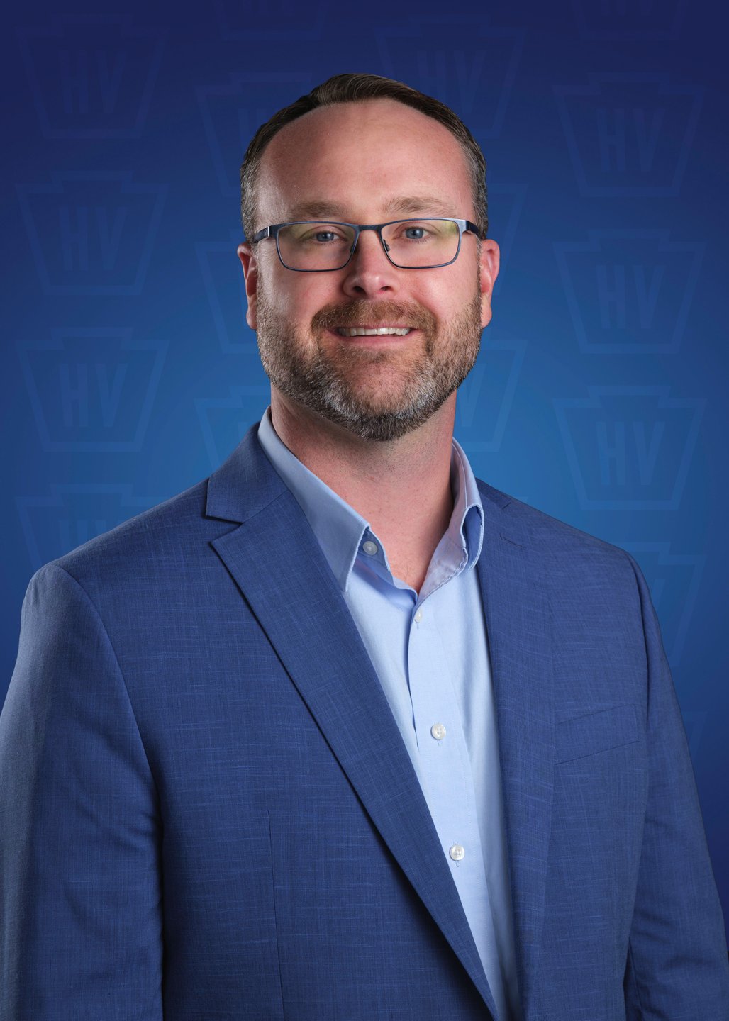A professional man in a blue suit and glasses smiles against a blue background with a subtle logo pattern, conveying a welcoming atmosphere.