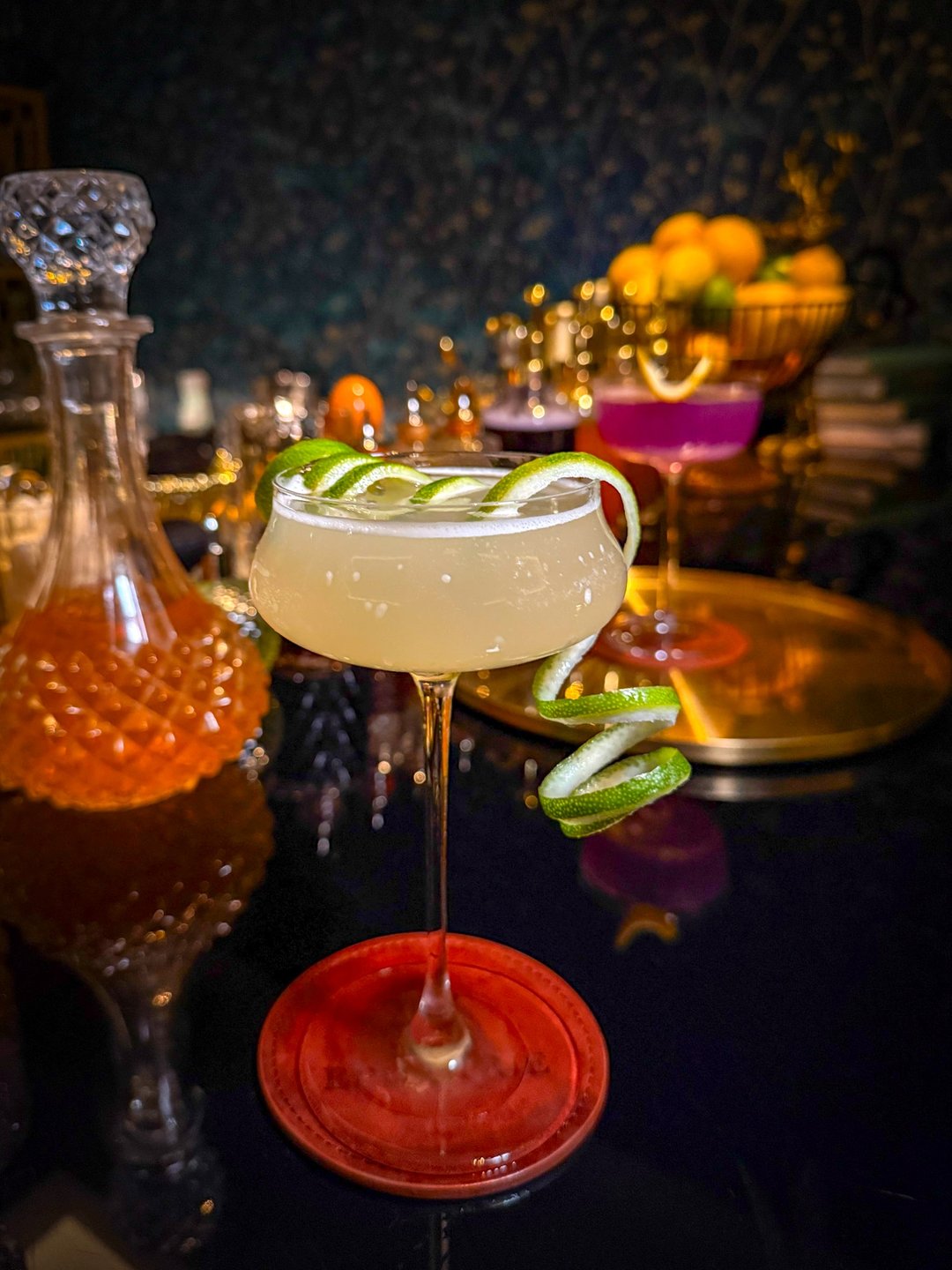 A stylish cocktail with a lime twist sits on a red coaster, surrounded by elegant glassware and vibrant garnishes in a dimly lit bar setting.