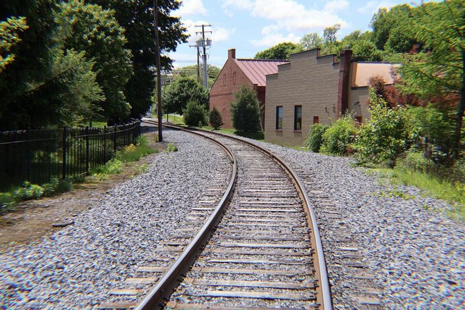 Curving railway tracks lined with gravel, flanked by greenery and buildings under a bright blue sky. Ideal for scenic exploration.
