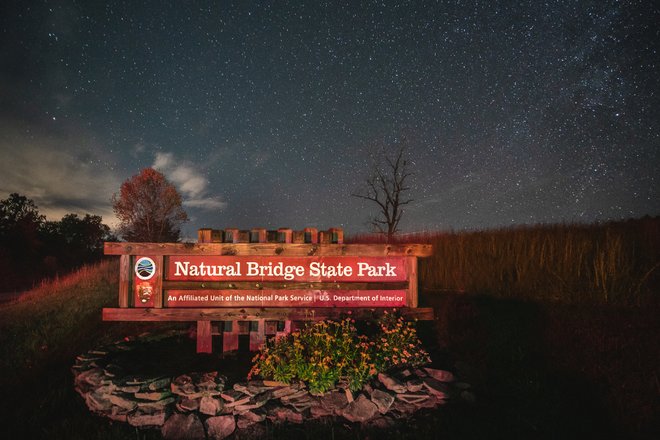 Sign for Natural Bridge State Park illuminated under a starry night sky, surrounded by grass and trees, promoting outdoor exploration.