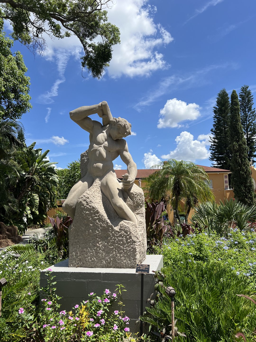 A stone sculpture of a muscular figure in a dynamic pose, surrounded by lush greenery and colorful flowers under a blue sky.