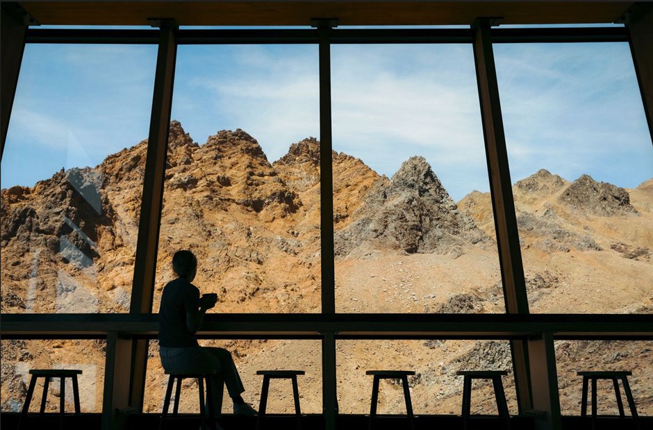 A person sits at a counter, gazing out large windows at rugged, rocky mountains under a clear blue sky. Stools are positioned nearby.