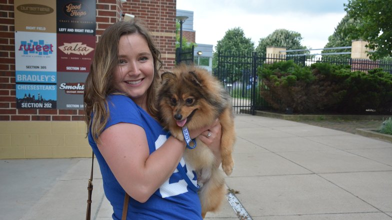 A woman holds a small, fluffy dog outside a building with signage. Green trees and a pathway are visible in the background.