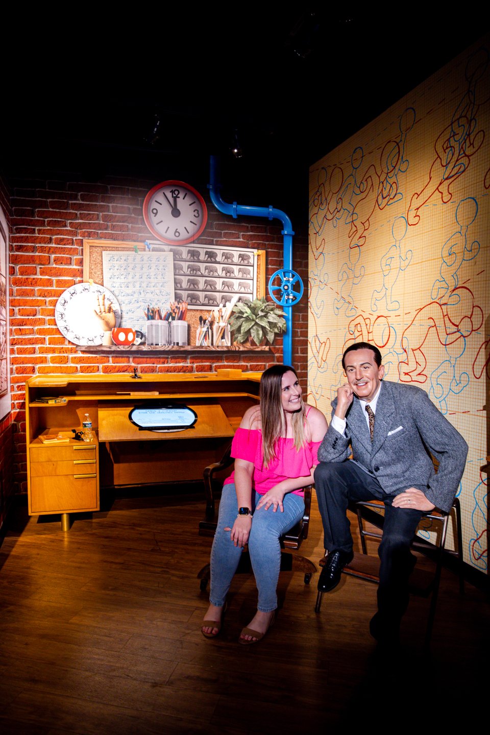 A woman in a pink top sits beside a lifelike figure in a gray suit, in a vintage-inspired room with brick walls and retro decor.