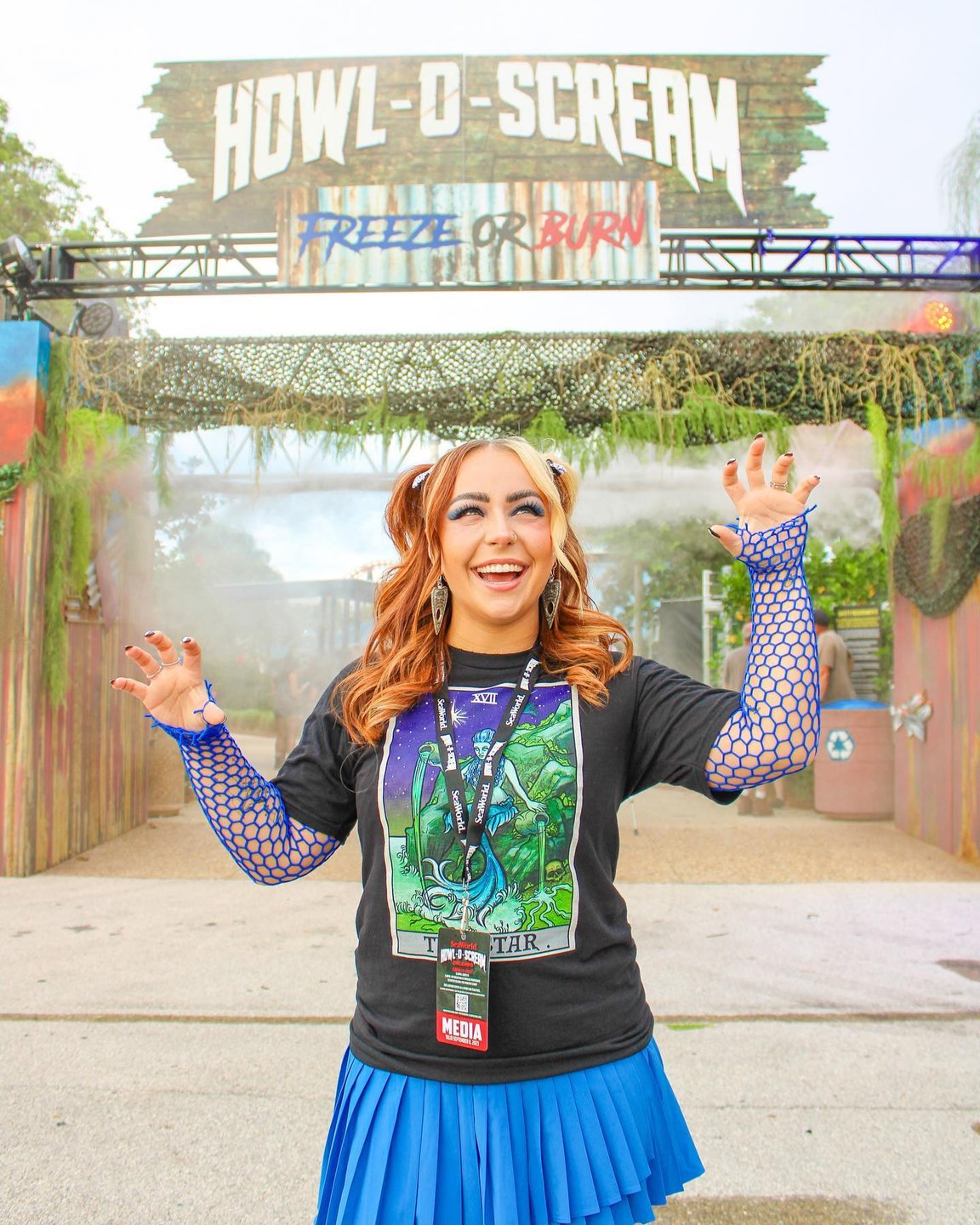 A young woman in a black graphic tee and blue skirt stands in front of a festive entrance sign for "Howl-O-Scream." The scene is lively, with fog effects and vibrant decorations.