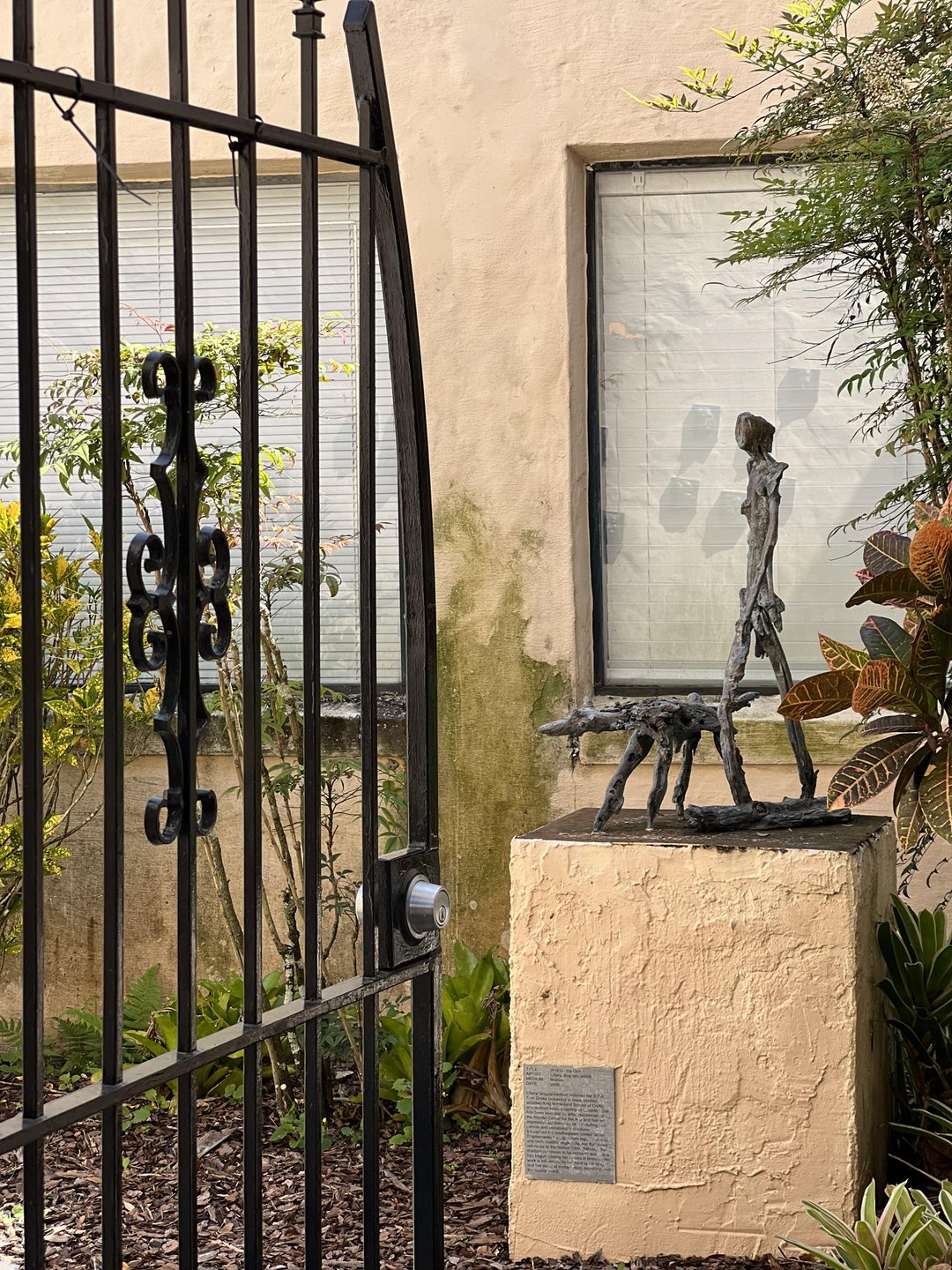 A wrought iron gate opens to reveal a bronze sculpture of a figure walking a dog, set against a textured wall and greenery.