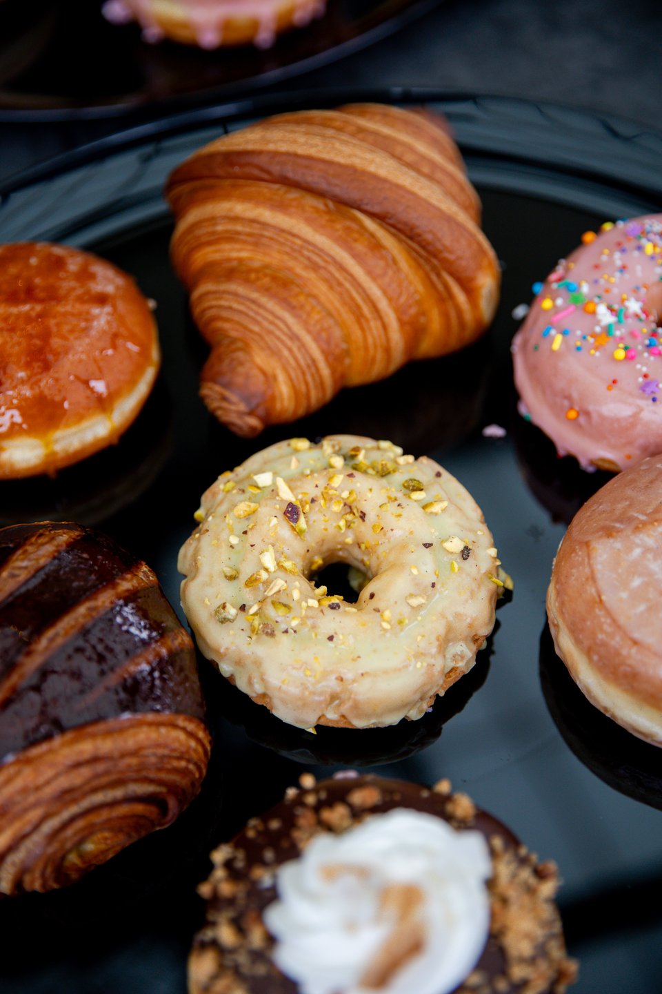 A variety of pastries on a black plate, including glazed donuts, a croissant, and a donut topped with sprinkles and nuts.