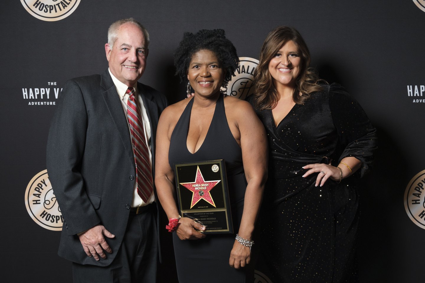 Three individuals pose together, smiling, in formal attire. One holds an award plaque featuring a star. The backdrop displays a logo for "The Happy Valley Adventure."