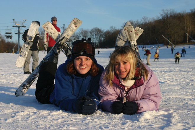 Two girls lie on the snow, smiling, with skis beside them. In the background, skiers navigate a slope under a clear sky.
