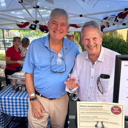 Two men smile at a food stall under a tent, surrounded by people enjoying a community event. A menu board displays local dishes.
