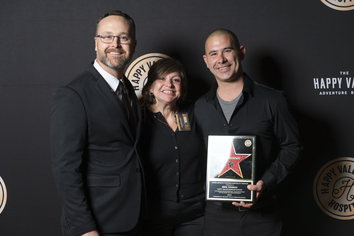 Three individuals stand together, smiling, holding an award plaque against a dark backdrop with a logo. The setting suggests a celebratory event.