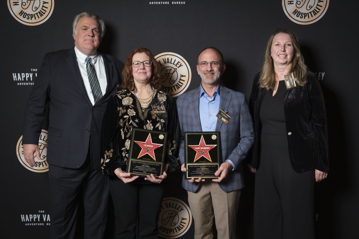 Four individuals stand together, holding awards on a stage with a dark backdrop featuring the "Happy Valley Adventure Bureau" logo.