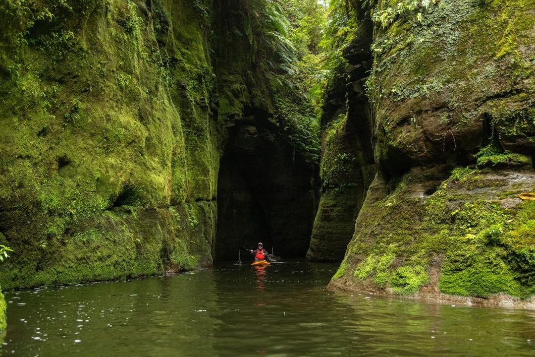 A kayaker navigates a narrow, lush green canyon with steep moss-covered walls, reflecting in the calm water below.