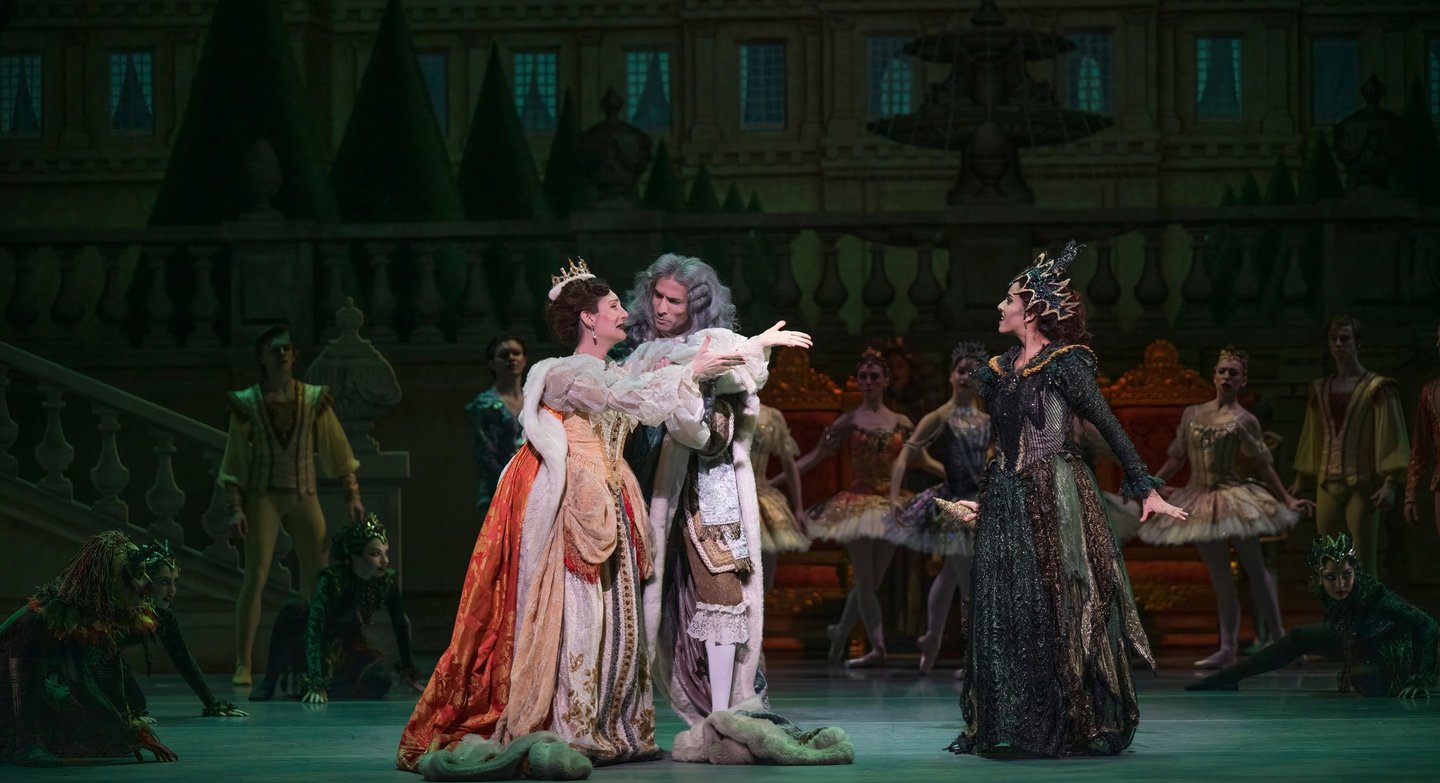 A theatrical performance featuring three elegantly dressed characters in a dramatic scene, surrounded by dancers in elaborate costumes.
