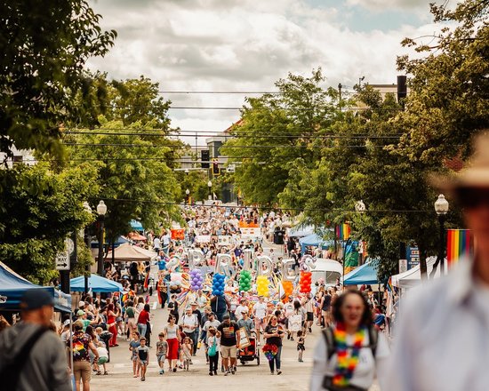 A vibrant street festival filled with people celebrating, colorful tents, and rainbow decorations. Trees line the street under a cloudy sky.