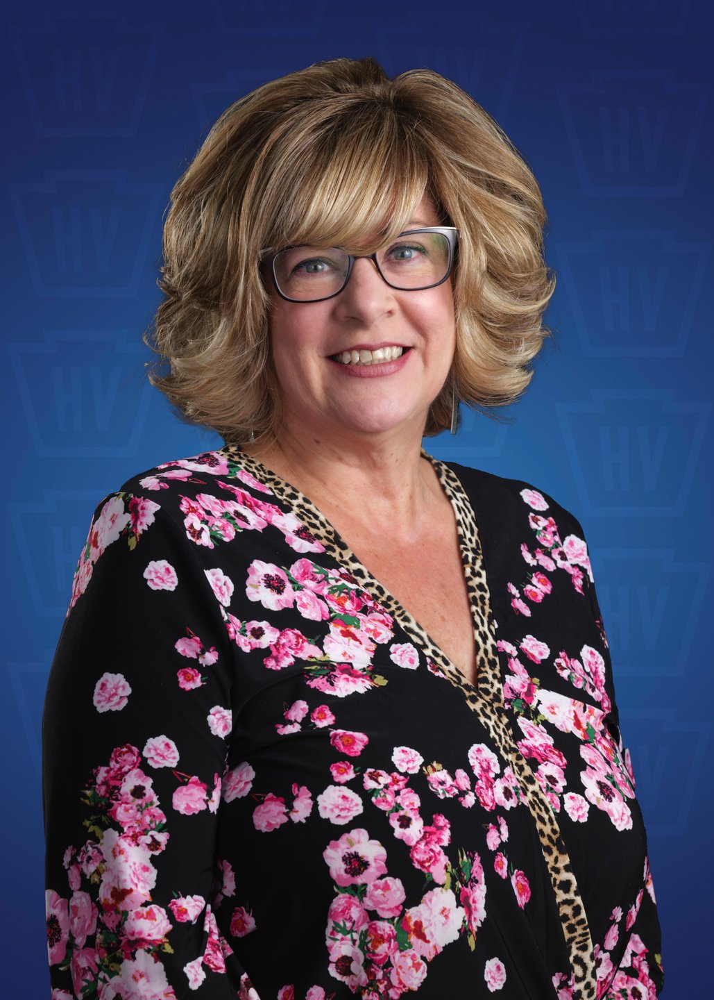 A woman with blonde, curly hair smiles, wearing a floral blouse against a blue background. Ideal for promotional travel content.