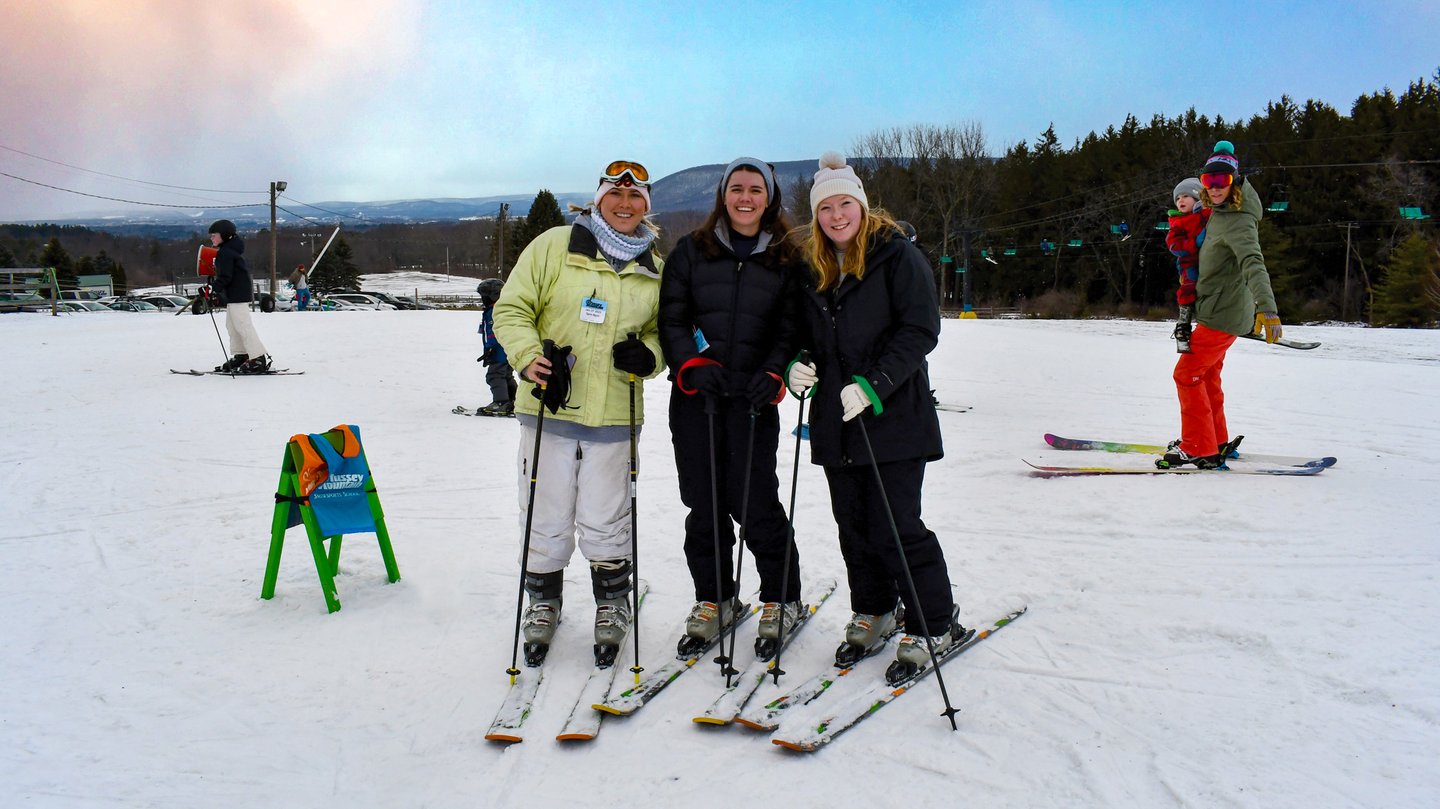 Three skiers pose on a snowy slope, equipped with ski poles and gear. In the background, other skiers enjoy the winter landscape.