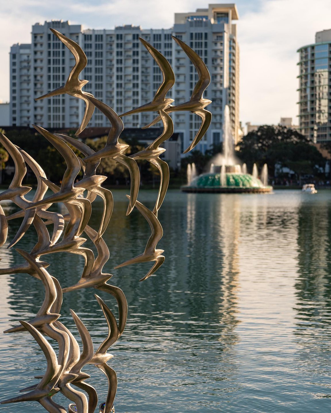 Sculpture of birds in flight stands by a serene lake, with a fountain and modern buildings in the background, creating a tranquil urban scene.