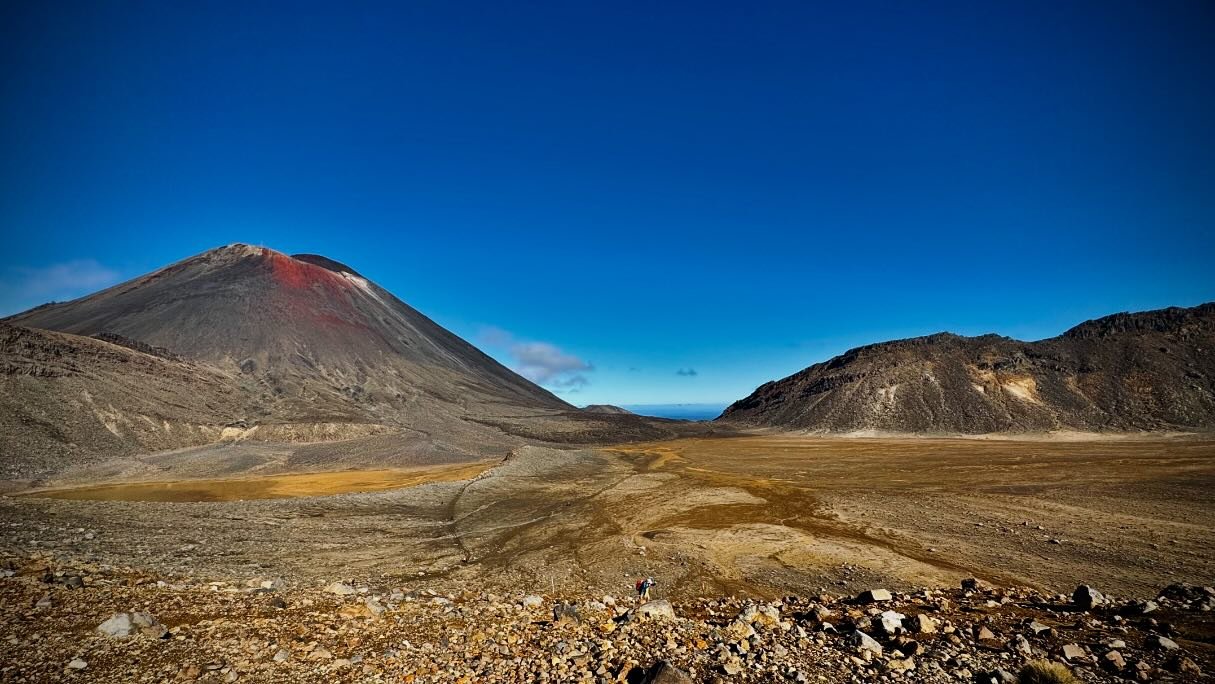 A vast volcanic landscape features a prominent cone-shaped volcano with a reddish summit, surrounded by rocky terrain and a clear blue sky.