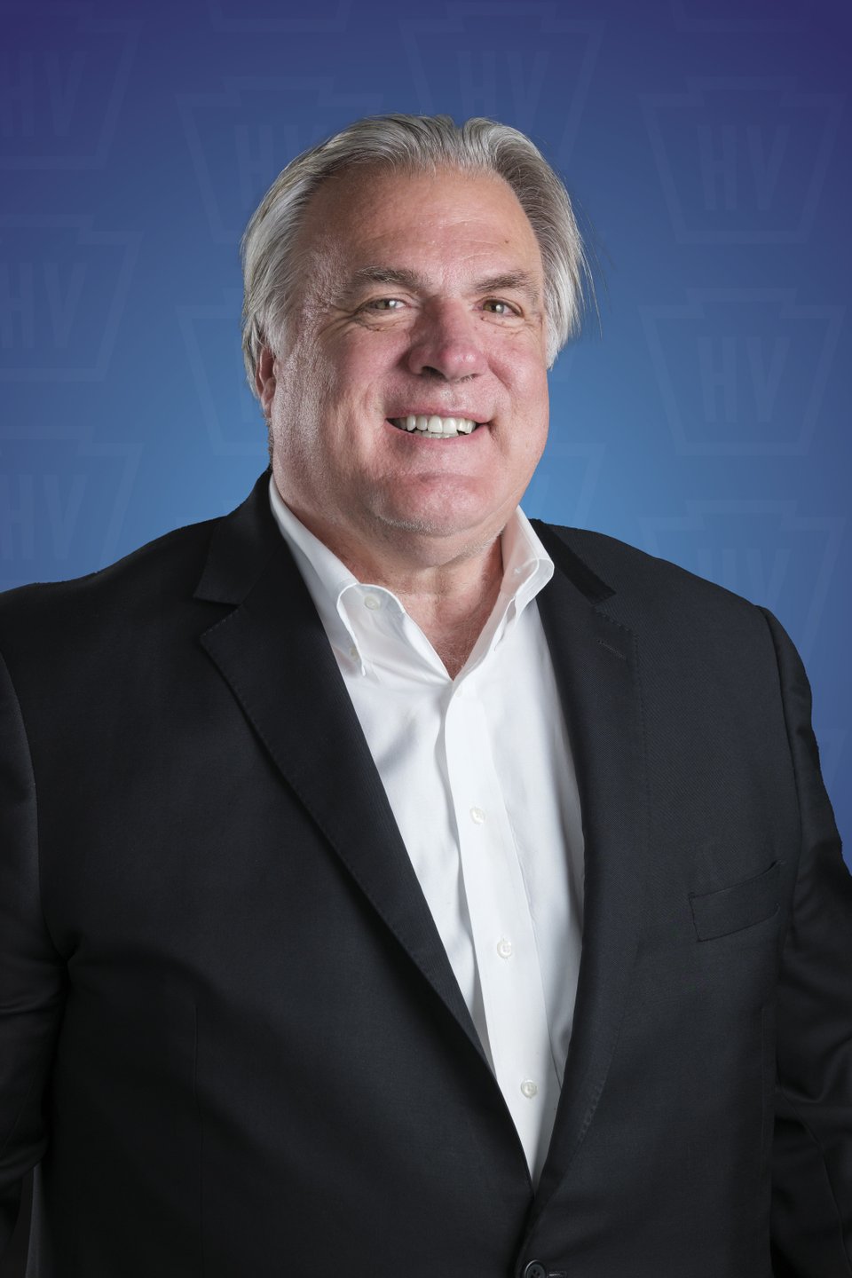A professional man in a black suit smiles against a blue background, promoting a welcoming atmosphere for business or networking events.