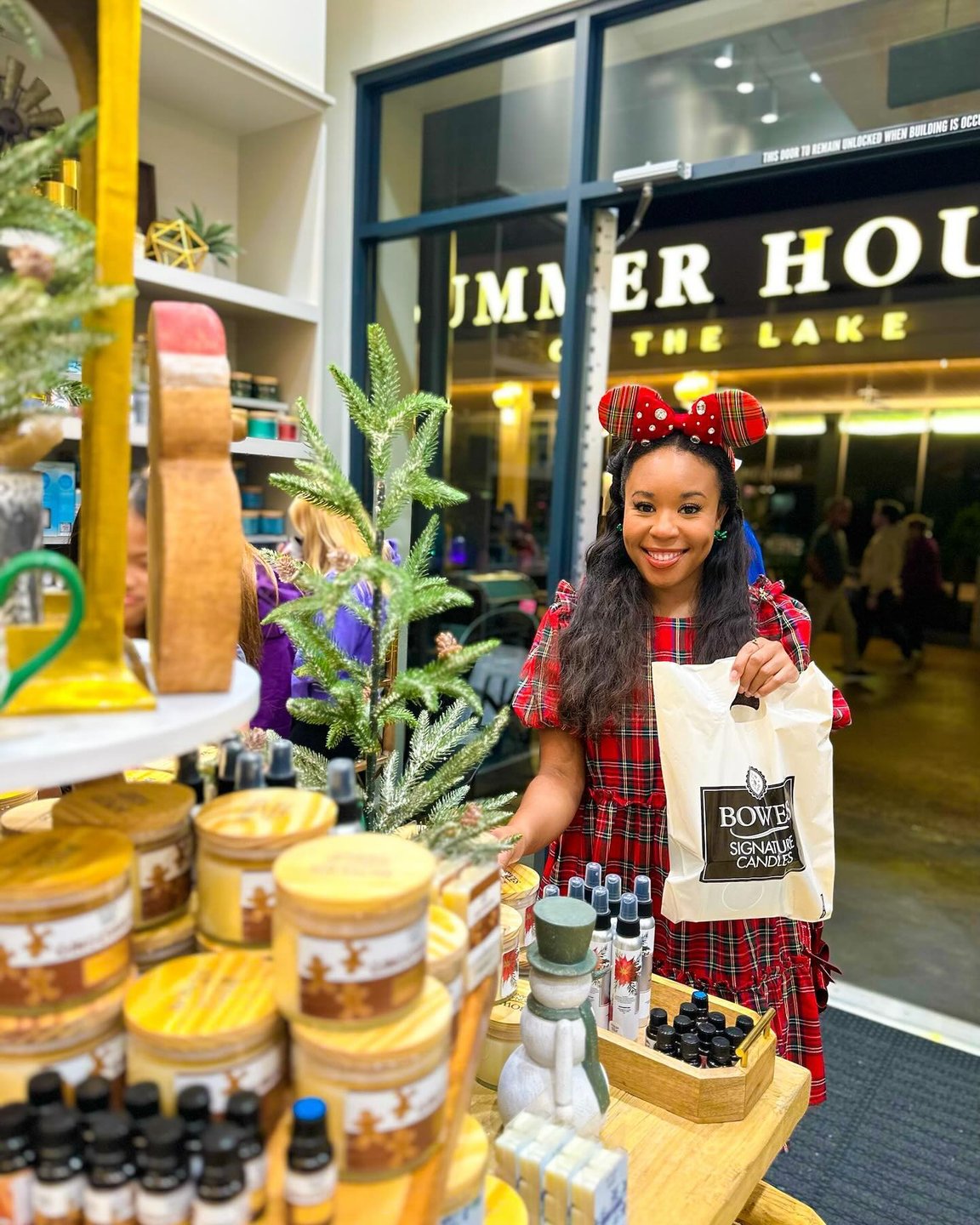 A woman in a festive outfit holds a shopping bag in a vibrant store filled with candles and skincare products. The background features a sign for "Summer House of the Lake."