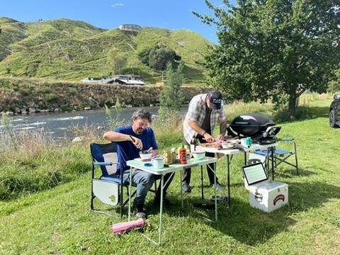 Two men prepare food at a picnic table by a river, surrounded by lush green hills and trees. A grill and cooler are nearby.