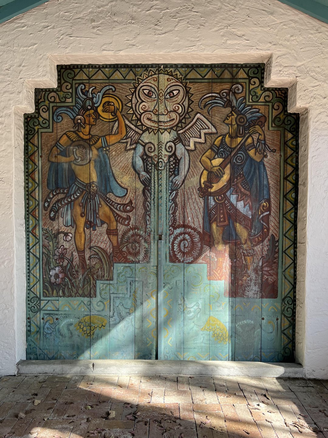 Mural depicting two figures in traditional attire, flanking a central sun motif on a weathered door. Sunlight casts shadows on the brick floor.