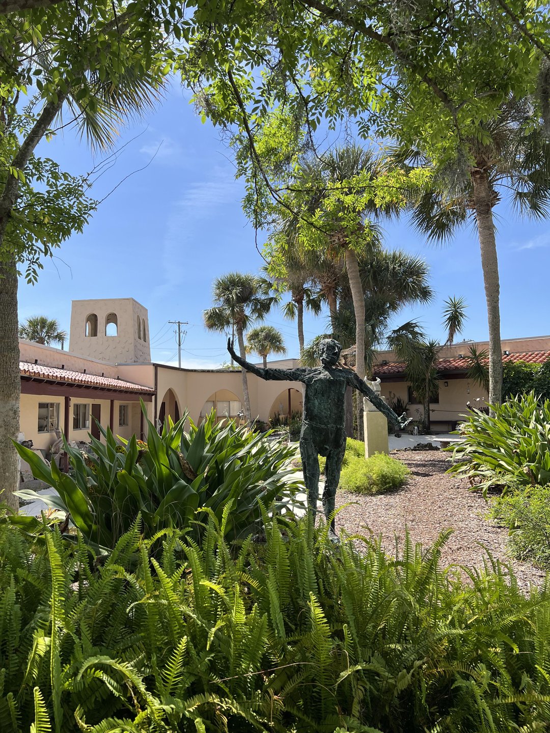 A bronze statue of a person stands amidst lush greenery and palm trees, with a building and blue sky in the background.