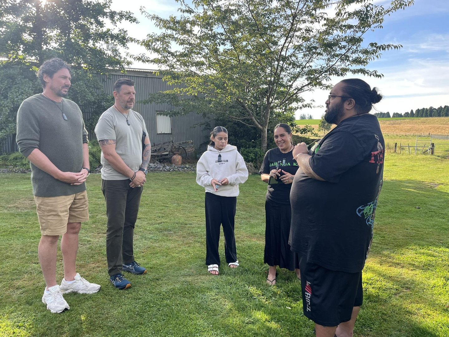 A group of five people stands in a grassy area, engaged in conversation. Trees and a field are visible in the background, suggesting a rural setting.